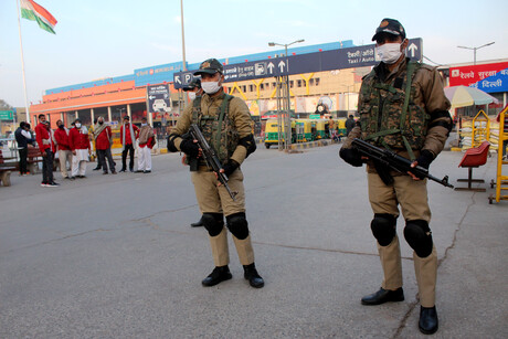 Zwei indische Polizisten stehen mit schusssicheren Westen, Knieschonern und Gewehren auf einem Platz; im Hintergrund sind Gebäude und weitere Personen zu erkennen