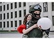 Eine junge Frau in einem roten Kleid umarmt einen Soldaten in Uniform, sie hält dabei einen Luftballon fest.