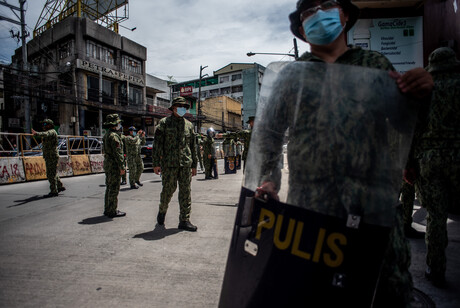 Philippinische Polizisten stehen über eine Straße versteut mit Masken dar, ein Polizist im rechten Vordergrund hält auch ein Schild vor dem Körper; im Hintergrund sind graue Häuser zu sehen